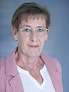 Eine Frau mit kurzen grauen Haaren, die eine Brille trägt, blickt freundlich in die Kamera. Sie trägt eine rosa Jacke über einem weißen Oberteil. Der Hintergrund ist einfarbig hellblau.