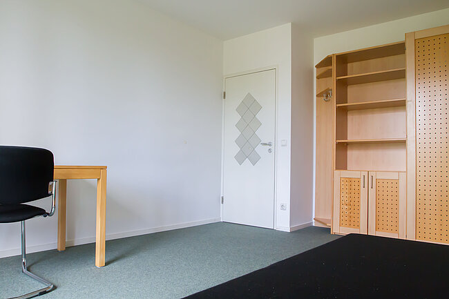 Ein leerer Raum mit weißer Wand, einer Tür mit diamantförmigem Muster, einem kleinen Tisch mit einem schwarzen Stuhl und einem Holzregal mit Schubladen und Fächern. Der Boden ist mit grauem Teppich ausgelegt.