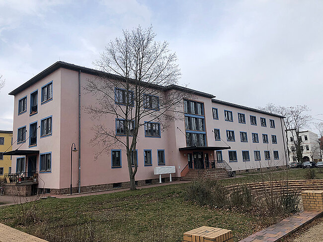 Außenansicht WH 2 Köthen; Foto: Thomas Faust Ein mehrstöckiges Gebäude mit hellrosa Fassade und vielen Fenstern. Vor dem Gebäude befindet sich ein kleiner, grasbewachsener Bereich mit einem Baum und einigen Sitzgelegenheiten. Der Himmel ist leicht bewölkt.