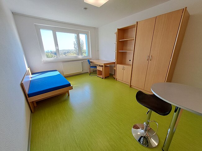 Ein einfaches, helles Zimmer mit grünem Bodenbelag. Es enthält ein Bett mit blauer Matratze, einen Schreibtisch mit Stuhl, einen großen Kleiderschrank und einen runden Tisch mit einem Hocker. Das Zimmer hat ein Fenster mit Blick auf Bäume und einen Heizkörper darunter.