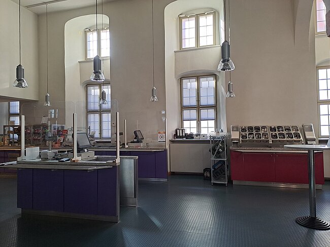 Ein Laborraum mit Arbeitsplätzen, Schränken und Fenstern. Der Raum ist mit mehreren Lampen ausgestattet, die von der Decke hängen. Es gibt Arbeitsflächen mit verschiedenen Geräten und Materialien. Der Boden ist mit Fliesen bedeckt und die Wände sind hell gestrichen.