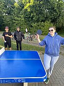 Drei Personen stehen in einem Park neben einem blauen Tischtennistisch. Eine Person hält einen Tischtennisschläger und macht eine spielerische Geste. Die anderen beiden Personen stehen im Hintergrund und lächeln. Im Hintergrund sind Fahrräder und Bäume zu sehen.