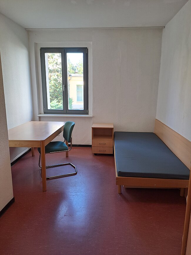 Ein einfaches, spartanisch eingerichtetes Zimmer mit einem Bett, einem Schreibtisch, einem Stuhl und einem kleinen Schrank. Das Zimmer hat ein Fenster, durch das Tageslicht einfällt. Der Boden ist mit rotem Linoleum ausgelegt, und die Wände sind weiß gestrichen.