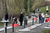 "Menschen spielen auf einem Mini-Golf-Platz im Freien. Der Platz ist mit Hindernissen und Kurven ausgestattet. Die Spieler sind auf einem gepflasterten Weg verteilt und konzentrieren sich auf das Spiel. Im Hintergrund sind Bäume und eine Wiese zu sehen."