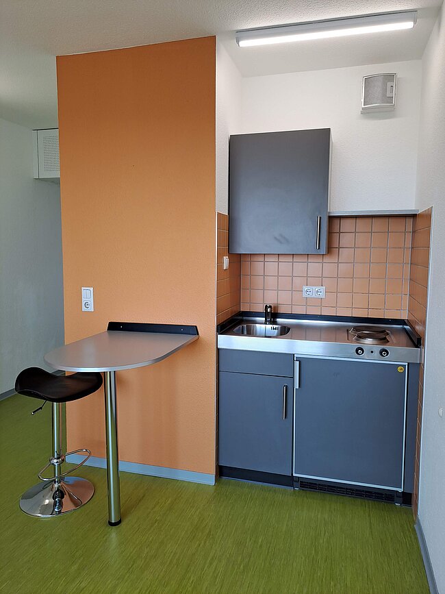 Ein kleiner Küchenbereich mit orangefarbener Wand, grauen Schränken, einem Spülbecken, einem Herd und einem Abzug. Daneben steht ein runder Tisch mit einem Hocker. Der Boden ist grün gefliest.