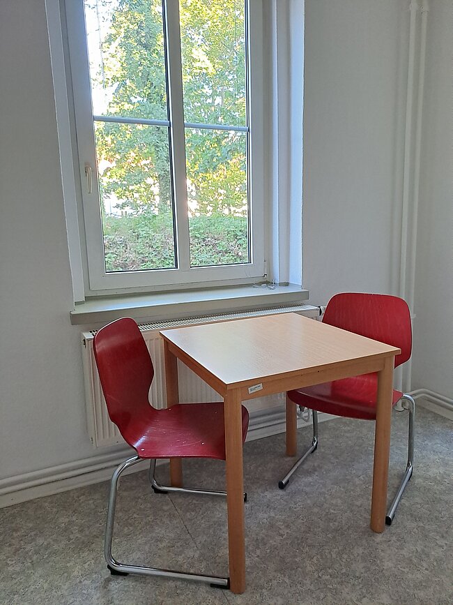 Ein kleiner Tisch mit zwei roten Stühlen steht vor einem Fenster. Durch das Fenster sieht man Bäume mit grünen Blättern. Der Raum hat helle Wände und einen grauen Teppichboden. Ein Heizkörper ist neben dem Fenster angebracht.