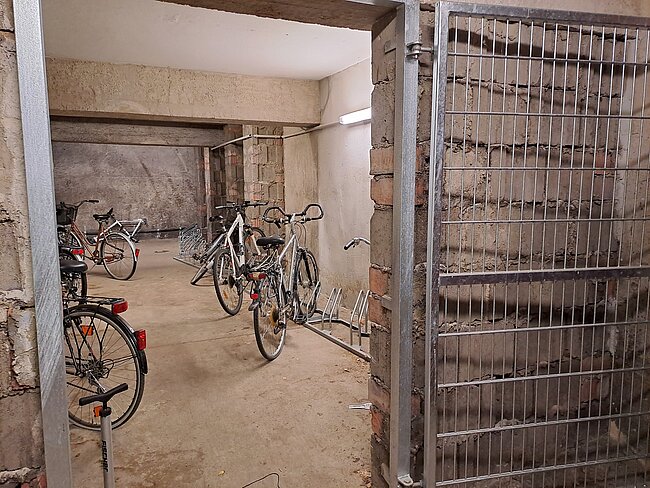"Ein Fahrradkeller mit mehreren Fahrrädern, die an Ständern befestigt sind. Der Raum hat Betonwände und einen Metallgitterverschluss an der Tür. Der Boden ist aus Beton und wirkt etwas schmutzig. Es gibt auch einen leeren Fahrradständer."