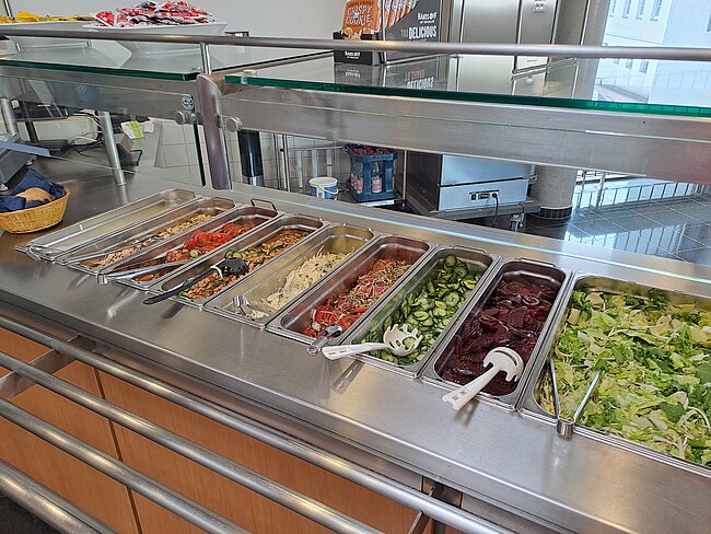 Ein Buffet mit verschiedenen Salaten und Beilagen in Edelstahlschüsseln. Es gibt grüne Salate, Nudelsalat, Bohnensalat und andere Gemüsegerichte. Servierlöffel liegen bereit. Die Anordnung ist sauber und übersichtlich.