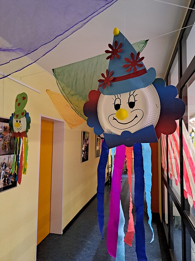 geschmückter Flur Ein bunter, handgemachter Luftballon mit einem lächelnden Gesicht, einem spitzen Hut und bunten Schleifen hängt von der Decke. Der Hintergrund zeigt einen Flur mit Bildern und farbigem Papierdekor.