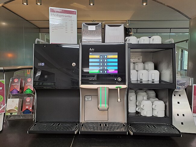Selbstbedienungs-Kaffeeautomat mit Touchscreen-Bedienfeld und farbigen Tasten zur Auswahl verschiedener Heißgetränke. Rechts daneben Regal mit Keramiktassen in verschiedenen Größen.