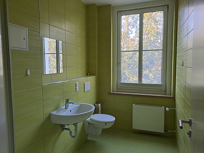 Ein modernes Badezimmer mit gelben Fliesen. Es gibt ein Waschbecken mit einem Spiegel darüber, eine Toilette und ein Fenster, durch das man Bäume sieht. An der Wand befindet sich ein Heizkörper. Der Raum wirkt hell und sauber.