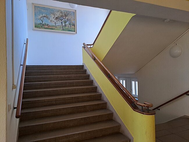 Ein Treppenaufgang mit Holzstufen und einem gelben Handlauf. An der Wand hängt ein Bild mit einer Landschaftsdarstellung. Die Treppe führt nach oben und hat ein Geländer auf beiden Seiten.