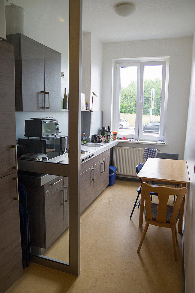 Küche in 4er-WG im Wohnheim Magdeburger Straße, Bernburg; Foto: Annegret Höhne Eine kleine, moderne Küche mit weißer Arbeitsplatte, grauen Schränken und einem Kühlschrank. Es gibt einen Tisch mit zwei Stühlen und ein Fenster, das auf eine Straße hinausgeht. Die Küche ist mit grundlegenden Küchengeräten ausgestattet, darunter ein Wasserkocher und eine Mikrowelle.
