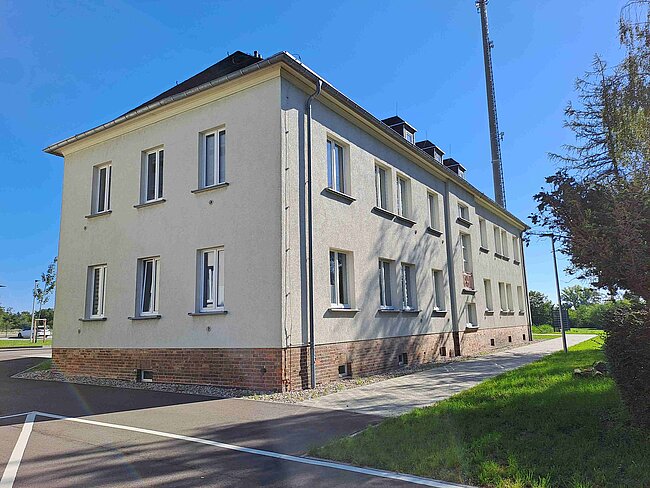 Ein zweistöckiges Gebäude mit hellbeiger Fassade und braunem Sockel. Es hat mehrere gleichmäßig angeordnete Fenster und befindet sich an einer Kreuzung mit einer asphaltierten Straße und einem grasbewachsenen Bereich. Der Himmel ist klar und blau.