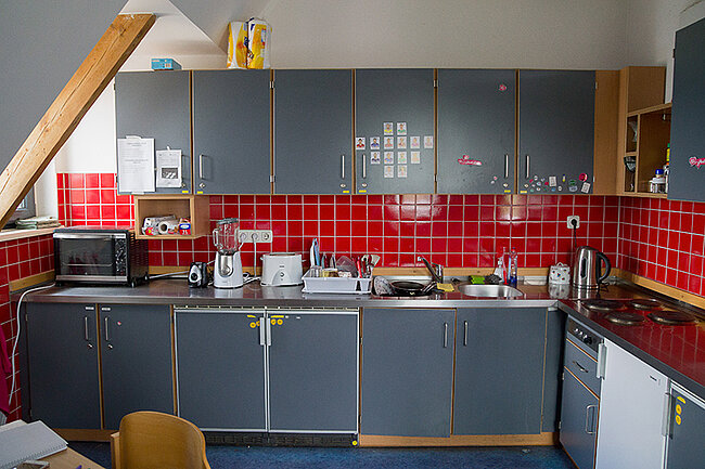 Küche in 10er-WG im Wohnheim Magdeburger Straße, Bernburg; Foto: Annegret Höhne Eine Küche mit grauen Schränken und roten Fliesen an der Wand. Auf der Arbeitsplatte stehen verschiedene Küchengeräte und Utensilien. Ein Mikrowellenherd ist links neben den Schränken platziert. Der Raum wirkt gut organisiert, aber etwas unordentlich.
