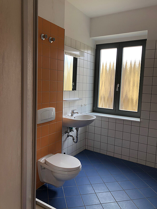 Bad WH 2 Köthen; Foto: Thomas Faust Ein kleines, sauberes Badezimmer mit weißem Waschbecken, einem weißen WC mit Wandmontage und einem Spiegel. Die Wände sind mit weißen Fliesen verkleidet, und es gibt ein Holzpaneel an einer Seite. Ein Fenster mit Blick auf Bäume lässt natürliches Licht herein. Der Boden ist mit blauen Fliesen bedeckt.