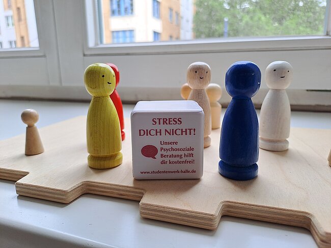 Holzfiguren auf einer Spielbahn mit farbigen Kleidungsstücken, dazu ein Anti-Stress-Würfel mit der Aufschrift „STRESS DICH NICHT!“ und Hinweis auf kostenfreie psychosoziale Beratung für Studierende.