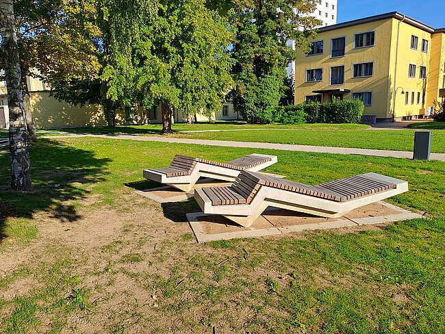 "Ein Park mit drei modernen Holzbänken, die in einer wellenförmigen Anordnung auf einem Betonsockel platziert sind. Im Hintergrund befindet sich ein gelbes Mehrfamilienhaus mit mehreren Fenstern. Der Bereich ist von Rasenflächen und Bäumen umgeben."