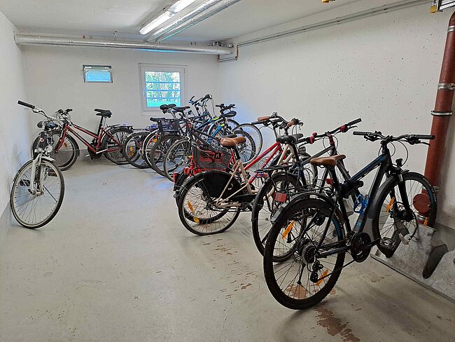 "Ein Fahrradkeller mit mehreren Fahrrädern, die an der Wand befestigt sind. Die Räder sind in verschiedenen Farben und Modellen vorhanden. Der Raum ist hell und hat ein Fenster. Der Boden ist aus Beton und wirkt sauber."