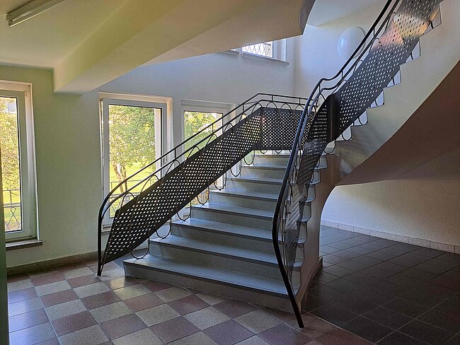 Ein Treppenhaus mit einer Wendeltreppe, die durch ein schwarzes Metallgeländer mit einem durchbrochenen Muster gesichert ist. Die Treppe führt nach oben und ist von Fenstern mit Tageslicht beleuchtet. Der Boden ist mit quadratischen Fliesen in Grau und Beige gestaltet.