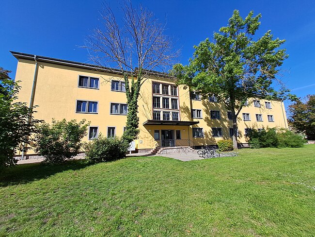 Ein gelbes Mehrfamilienhaus mit mehreren Fenstern und einem zentralen Eingang. Vor dem Gebäude befindet sich ein gepflegter Rasen mit einigen Büschen und Bäumen. Der Himmel ist klar und blau.