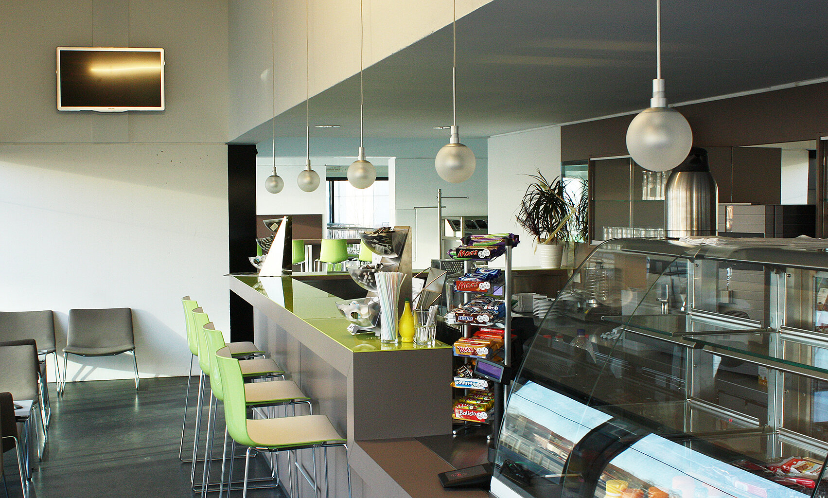 Cafebar Heidemensa; Foto: Thomas Faust Ein moderner Café- oder Restaurantbereich mit einer Theke, an der verschiedene Snacks und Getränke angeboten werden. Es gibt Hocker zum Sitzen und eine Küche im Hintergrund. Die Einrichtung ist hell und zeitgemäß mit großen Fenstern, die viel Licht hereinlassen.