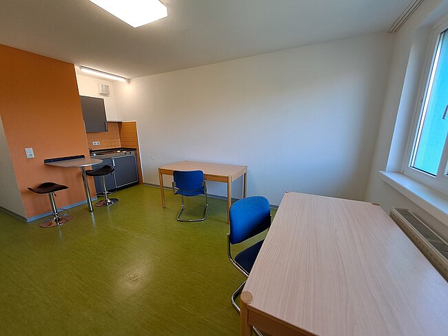 Ein kleiner, sauberer Raum mit grünem Boden. Es gibt einen Tisch mit zwei blauen Stühlen, einen hohen Tisch mit zwei Hockern und eine Küchenecke mit einem kleinen Kühlschrank. Ein Fenster lässt Tageslicht herein. Die Wände sind weiß und orange.