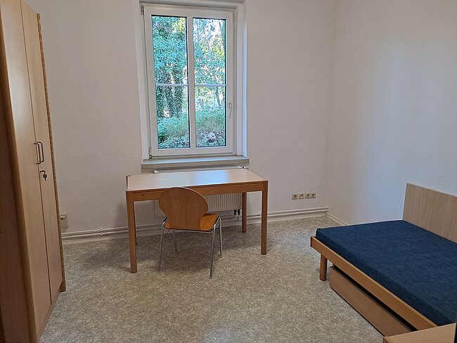 Ein einfaches Zimmer mit einem Schreibtisch, einem Stuhl und einem Bett. Der Schreibtisch steht vor einem Fenster mit Blick auf Bäume. Die Wände sind weiß und der Boden ist mit grauem Teppichboden bedeckt. Die Tür ist links im Bild zu sehen.