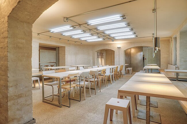 Speisesaal (© Patrick Müßiggang) Ein moderner, heller Raum mit mehreren Tischen und Stühlen. Die Tische sind in Reihen angeordnet und haben eine schlichte, minimalistische Gestaltung. Die Wände sind aus hellem Stein, und die Decke ist mit mehreren Reihen von Leuchtstoffröhren beleuchtet. Der Boden ist mit hellen Fliesen bedeckt.