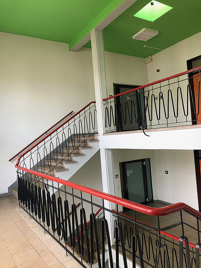 Treppenhaus WH 2 Köthen; Foto: Thomas Faust Ein Treppenhaus mit zwei Etagen, das durch ein Geländer mit roten Handläufen und schwarzen Metallstäben begrenzt ist. Die Decke ist grün gestrichen, und es gibt Türen auf beiden Ebenen. Die Wände sind weiß, und der Boden besteht aus Fliesen.