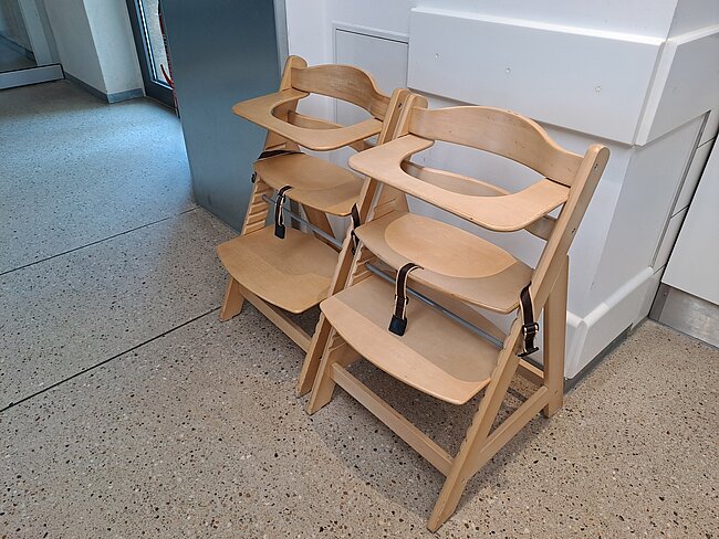 Zwei Hochstühle aus hellem Holz mit Trittstufe und Armlehnen.