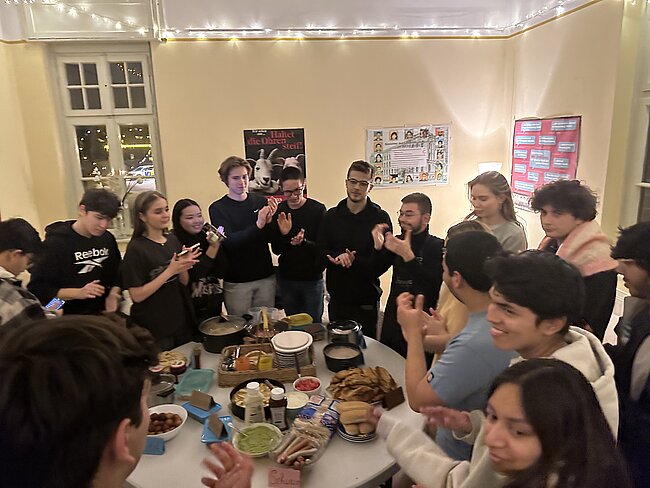 Multi-Kulti-Kochen im Wohnheim Eine Gruppe von Menschen steht um einen Tisch mit verschiedenen Speisen und Getränken. Sie scheinen sich zu unterhalten und zu feiern. Die Atmosphäre wirkt fröhlich und gesellig.