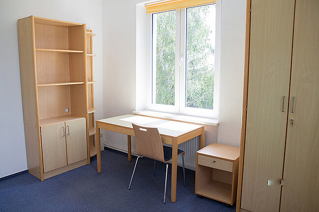 Zimmer in 5er-WG im Wohnheim Magdeburger Straße, Bernburg; Foto: Annegret Höhne Ein einfaches Zimmer mit einem Schreibtisch, zwei Stühlen, einem hohen Regal mit Schubladen und einem kleinen Beistelltisch. Ein Fenster mit Vorhang lässt Tageslicht herein. Die Wände sind weiß und der Boden ist mit blauen Teppichfliesen bedeckt.