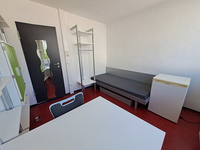 Ein einfaches Zimmer mit einem Bett, einem Schreibtisch, einem Stuhl, einem Kleiderschrank und einem Kühlschrank. Die Wände sind weiß, und der Boden ist mit rotem Teppich ausgelegt. Es gibt auch einen Kleiderständer und eine Tür mit einem bunten Poster daran.