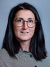 Eine Frau mit langen dunklen Haaren, die einen Pony trägt, und einer Brille. Sie trägt ein gemustertes Hemd mit einem geometrischen Muster. Sie lächelt leicht und blickt direkt in die Kamera.