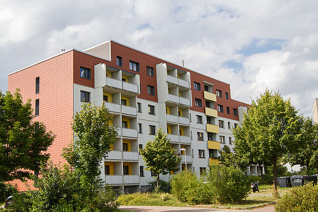 Ein modernes Mehrfamilienhaus mit mehreren Stockwerken, das in hellen Farben gestrichen ist. Die Fassade ist mit roten und weißen Panelen gestaltet. Vor dem Gebäude befinden sich Bäume und Büsche, die eine grüne Umgebung schaffen. Der Himmel ist teilweise bewölkt.