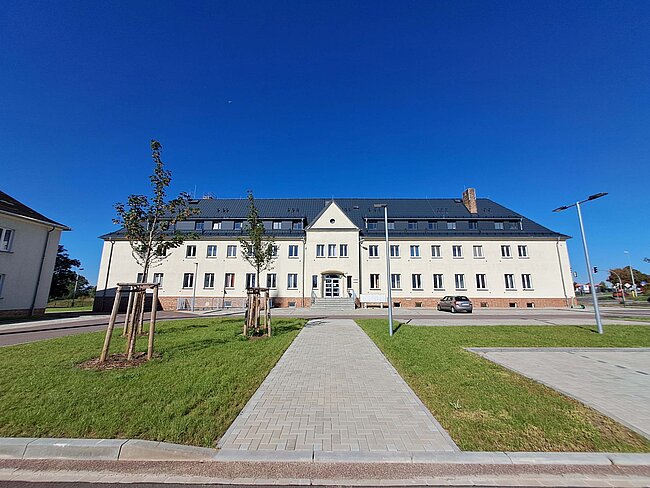 Ein großes, weißes Gebäude mit einem zentralen Eingang und einem gepflasterten Weg, der darauf zuführt. Das Gebäude hat zwei Stockwerke und ein schwarzes Dach. Vor dem Gebäude befinden sich junge Bäume und eine gut gepflegte Rasenfläche. Der Himmel ist klar und blau.