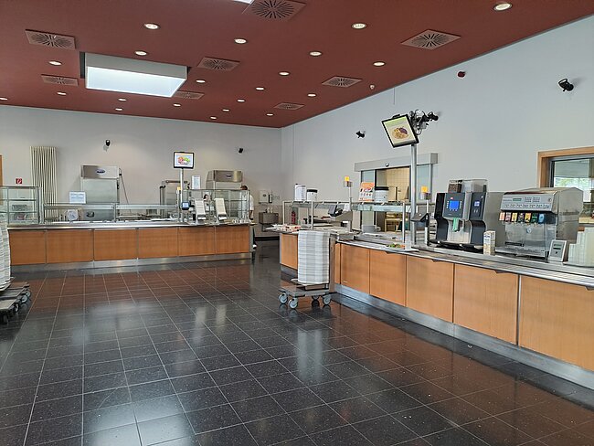 Ausgabebereich Ein sauberer, moderner Küchenbereich mit Arbeitsflächen, Geräten und Geschirrspülern. Es gibt Theken mit Kassen und Kaffeemaschinen. Der Boden ist mit dunklen Fliesen belegt, und die Decke hat rote Paneele mit eingebauten Lampen. Es sind keine Personen zu sehen.