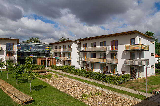 Moderne Wohnanlage mit mehreren Gebäuden, die durch einen gepflegten Garten mit Wegen und Bäumen verbunden sind. Die Häuser haben Balkone und große Fenster. Der Himmel ist bewölkt, aber es scheint sonnig zu sein. Die Anlage wirkt ruhig und einladend.
