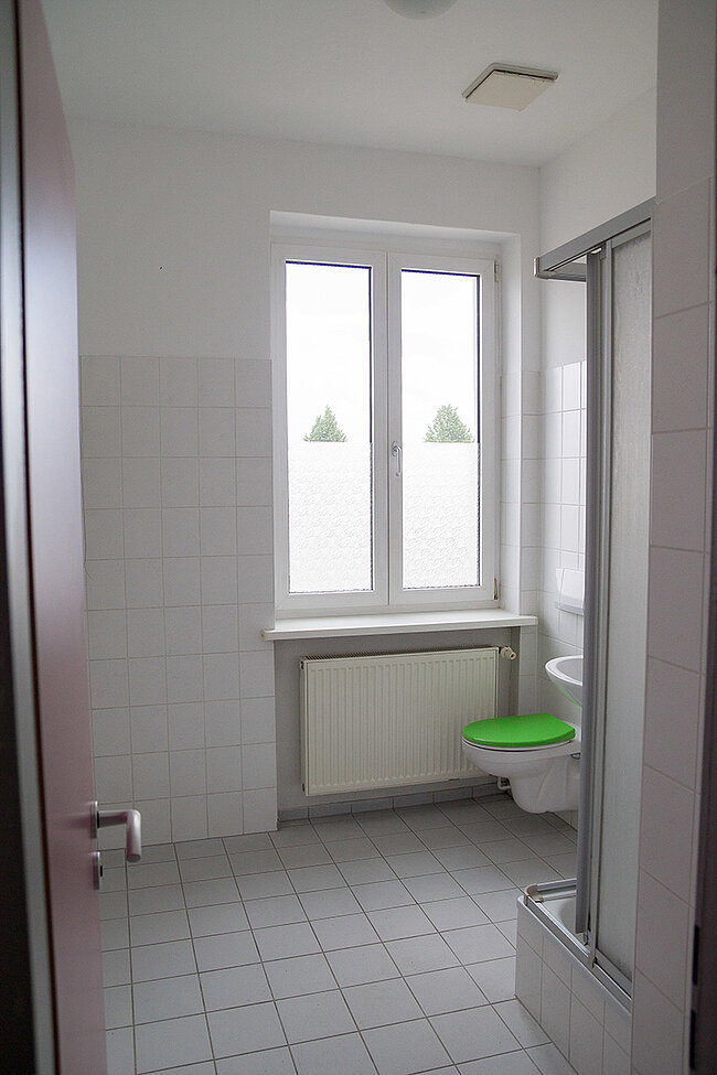 Bad in 4er-WG im Wohnheim Magdeburger Straße, Bernburg; Foto: Annegret Höhne Ein weiß gefliestes Badezimmer mit einem grünen Toilettensitz. Es gibt ein Fenster mit zwei Flügeln, darunter ein weißes Waschbecken und ein Heizkörper. Rechts befindet sich eine Duschkabine mit einer Glastür. Der Boden ist ebenfalls weiß gefliest.