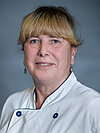 Eine Frau mit blonden Haaren und blauen Augen trägt eine weiße Kochjacke mit blauen Knöpfen. Sie lächelt leicht und hat Ohrringe an. Der Hintergrund ist grau und unauffällig.