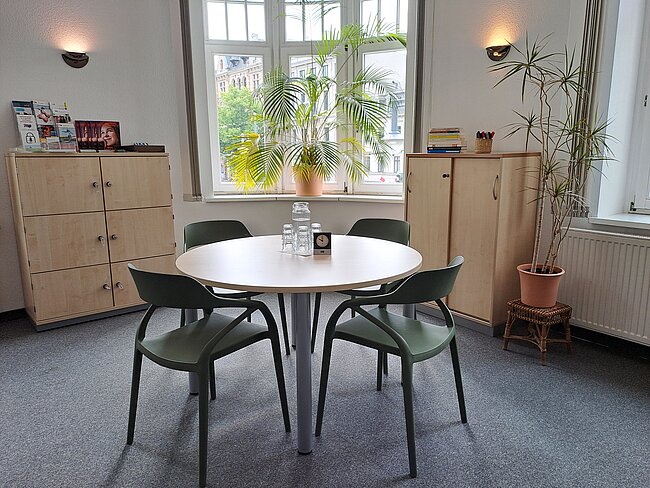 Heller, moderner runder Besprechungstisch mit vier Stühlen in einem hellen Raum. Im Hintergrund große Fenster mit Blick nach draußen, zwei Pflanzen, Regale mit Ordnern und Bücher. Tischmitte: kleine Dose und Stiftehalter.