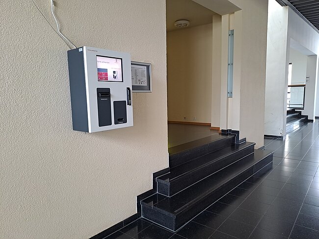 Aufwerter Ein Treppenaufgang mit einem schwarzen Handlauf. An der Wand links neben den Stufen ist ein grauer Kasten mit einem Bildschirm und Tasten befestigt. Der Boden ist mit dunklen Fliesen verlegt. Der Hintergrund zeigt einen hellen Flur mit einer Tür.