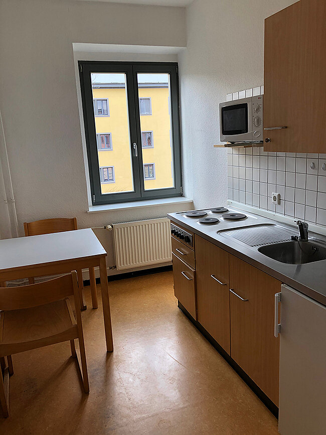 Küche WH 2 Köthen; Foto: Thomas Faust Eine kleine Küche mit einem Fenster, das auf ein gelbes Gebäude zeigt. Es gibt einen Herd mit zwei Kochplatten, eine Spüle und einen Mikrowellenofen. Daneben steht ein Tisch mit zwei Stühlen. Die Küche hat helle Holzmöbel und geflieste Wände.