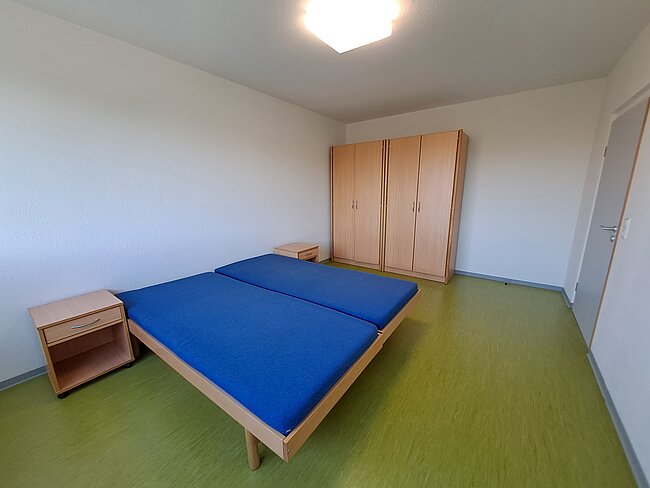 Ein einfaches Zimmer mit zwei schmalen Betten, die mit blauen Matratzen bezogen sind. An der Wand steht ein großer Kleiderschrank aus hellholz. Der Boden ist mit grünem Teppichboden bedeckt. Es gibt zwei kleine Nachttische neben den Betten.