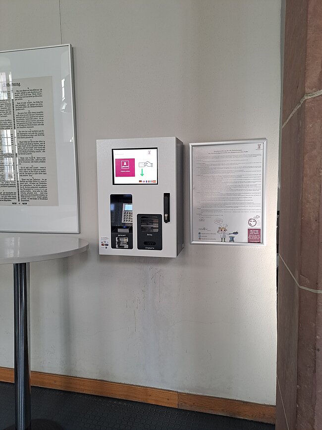 Ein Bildschirm mit einer Handdesinfektionsstation und Anweisungen zur Handhygiene. Links daneben ist eine Bibel in einem Ständer. Rechts befindet sich ein Informationsblatt mit weiteren Hinweisen. Die Station ist in einem Raum mit weißer Wand und Holzboden aufgestellt.