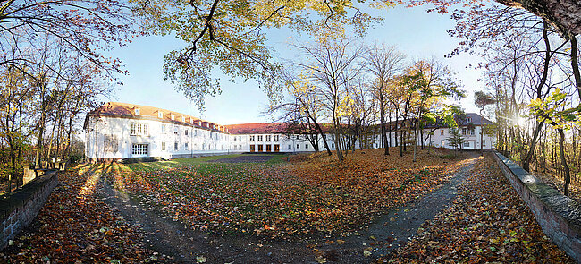 Ein großes weißes Gebäude mit rotem Dach steht in einem parkähnlichen Gelände mit vielen Bäumen. Der Boden ist mit herabgefallenen Blättern bedeckt. Die Szene wirkt friedlich und ist von Natur umgeben.