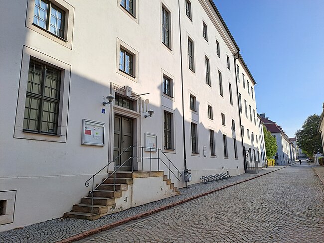 Ein weißes Gebäude mit mehreren Fenstern und einem Eingang mit einer Treppe. Der Eingang hat eine Rampe und ein Schild. Die Straße davor ist mit Kopfsteinpflaster gepflastert und verläuft geradeaus. Die Fassade des Gebäudes ist schlicht und modern.