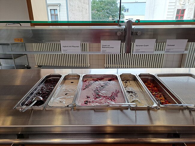 Desserttheke Ein Buffet mit verschiedenen Joghurt- und Quarksorten. Von links nach rechts: Joghurt mit Früchten, Quark mit Beeren, Kirschquark und Quark mit Obst. Die Behälter sind aus Edelstahl und stehen auf einer glatten Metalloberfläche.