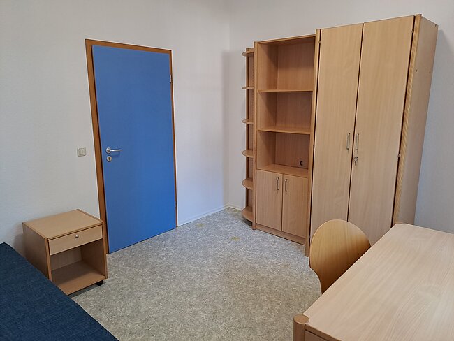 Ein einfacher Raum mit einem blauen Schrank, einem Bücherregal, einem Schreibtisch mit Stuhl und einem kleinen Nachttisch. Der Boden ist mit grauem Teppich bedeckt. Die Wände sind weiß und es gibt eine Tür mit einem blauen Panel.